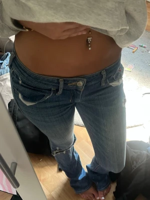 Blå jeans från Hollister - Säljer ett par blå jeans från Hollister med slitningar och snygga detaljer på bakfickorna. Jeansen har låg midja och klassisk femficksdesign. Perfekta för dig som gillar en avslappnad och trendig stil.