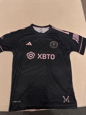 Svart Inter Miami Messi tröja - Säljer en svart Inter Miami matchtröja med rosa detaljer och Messi 10 på ryggen. Tröjan har korta ärmar, rund hals och tryckta loggor samt sponsortryck framtill. Perfekt för fotbollsfans som vill stötta sitt lag med stil.
