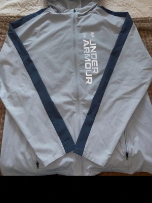 Ljusgrå vindjacka från Under Armour - Snygg ljusgrå vindjacka från Under Armour med mörkblå detaljer längs ärmarna och dragkedja framtill. Jackan har huva, två sidofickor med dragkedja och logga tryckt i vitt på bröstet. Perfekt för blåsiga dagar och sportiga outfits.