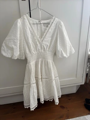 Vit kort klänning från By Malina - Superfin vit kort klänning från By Malina med v-ringning, puffärmar och volangdetaljer. Klänningen har broderade spetskanter och ett diskret prickigt mönster. Perfekt för sommarens alla tillfällen, till konfa, student eller bara en fin middag❤️Har bara använt 1 gång på min student🩷