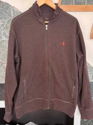 Brun zip-tröja från Ralph Lauren - Mörkbrun tröja med dragkedja från Ralph Lauren. Klassisk modell med dragkedja, två fickor framtill och den ikoniska orangea loggan broderad på bröstet. Tillverkad i mjukt bomullsmaterial som är skönt att ha på sig. Perfekt för en avslappnad och stilren look.