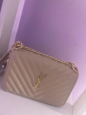 Beige YSL quiltad handväska - Snygg beige quiltad handväska från YSL med guldfärgad kedjerem och ikonisk YSL-logga framtill. Väskan har ett stilrent mönster i v-form och är tillverkad i slätt, glansigt material. Perfekt accessoar för att lyfta din outfit.