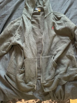 Svart hoodie från Polo Ralph Lauren - Säljer en svart hoodie från Polo Ralph Lauren i storlek M. Hoodien har dragkedja, två stora fickor framtill och en liten röd logga på bröstet. Tillverkad i mjukt och bekvämt material, perfekt för chill dagar. Klassisk design med huva.