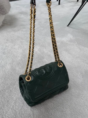 Tory Burch väska  - Säljer denna superfina höst väska i mörkgrönt från Tory Burch med guld detaljer. Den är i fint skick. Väskan går både att ha över axeln men även med långt band. Den har många olika fack att sortera upp sina saker i. 