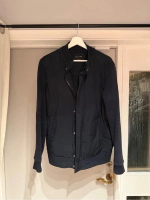 Mörkblå bomberjacka med tryckknappar - Stilren mörkblå cardigan med tryckknappar framtill och ribbade muddar vid ärmslut och nederkant. Jackan har en minimalistisk design utan synliga loggor och är tillverkad i ett slätt tyg. Perfekt för dig som gillar enkel och clean stil.