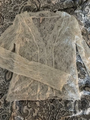Vit spetsblus från Nelly - Säljer en superfin vit spetsblus från Nelly med v-ringning och långa ärmar. Blusen är helt i spets med blommigt mönster och genomskinlig look, perfekt för att styla lager på lager. 
