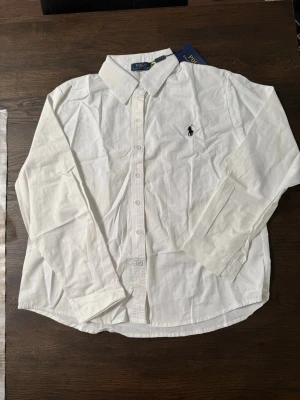 Vit skjorta från Polo Ralph Lauren - Klassisk vit skjorta från Polo Ralph Lauren med svart broderad logga på bröstet. Skjortan har lång ärm, knappar framtill och en tidlös krage. Tillverkad i mjuk bomull som känns skön mot huden. Perfekt för en clean och stilren look.