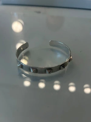 Silvrigt armband med nitar - Snyggt silvrigt armband i metall med pyramidformade nitar runt hela bandet. Armbandet är stelt och har en öppen design som gör det enkelt att ta på och av. Perfekt för dig som gillar en edgy och modern stil.