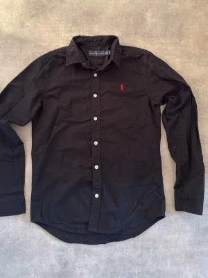 Svart skjorta från Ralph Lauren - Klassisk svart skjorta från Ralph Lauren med vit knappstängning och röd broderad logga på bröstet. Skjortan har lång ärm, krage och är tillverkad i bomull. Perfekt för en clean och stilren look.