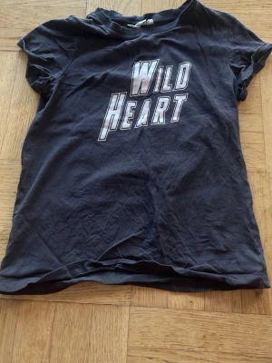 Svart t-shirt Wild Heart KappAhl - Cool svart t-shirt från KappAhl med trycket 'Wild Heart' i stora bokstäver på bröstet. Klassisk passform med korta ärmar och rund hals. Perfekt till jeans eller shorts för en avslappnad look.