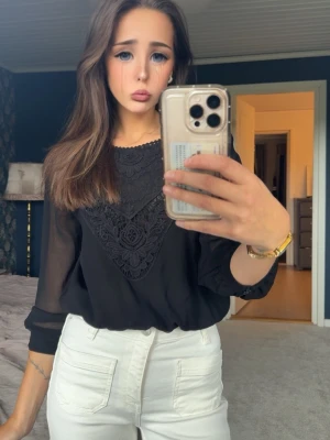 Svart blus med spetsdetaljer - Snygg svart blus med transparenta långärmar och dekorativ spets framtill. Blusen har rund halsringning och ett luftigt, lätt material som ger en elegant look. Perfekt att matcha med både jeans och kjol för en trendig outfit.