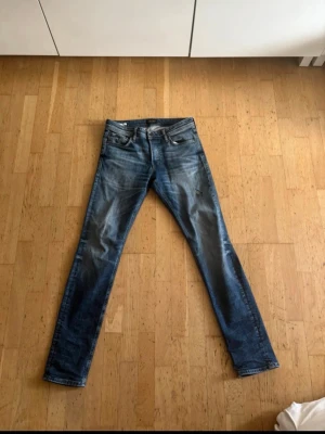 Jack & Jones  - Säljer ett par blå jeans med tvättad look och ett tydligt hål på ena benet. Jeansen har klassisk femficksdesign och raka ben. Perfekta för dig som gillar en avslappnad och trendig stil.