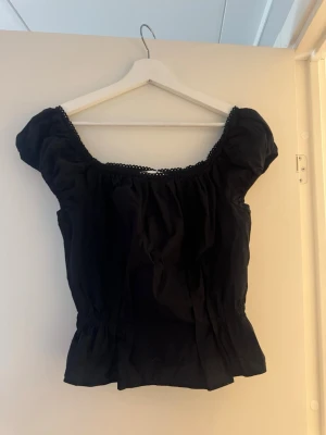 Svart puffärm blus med resår - Söt svart blus med puffärmar och resår i midjan för en smickrande passform. Toppen har en rundad halsringning med liten spetskant och är perfekt för dig som gillar en feminin och trendig stil.