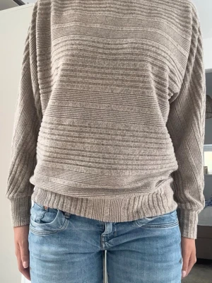 Beige ribbad stickad tröja - Mysig beige stickad tröja med horisontella ribbade ränder. Tröjan har rund halsringning, långa ärmar och breda muddar vid ärmslut och nederkant. Perfekt för lager på lager och enkel att matcha med jeans.