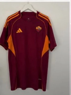 Vinröd AS Roma träningströja Adidas - Snygg vinröd träningströja från Adidas med AS Roma-logga på bröstet. Tröjan har orange detaljer på axlar och sidor samt klassiska Adidas-ränder. Tillverkad i lätt och funktionellt material, perfekt för fotbollsträning.