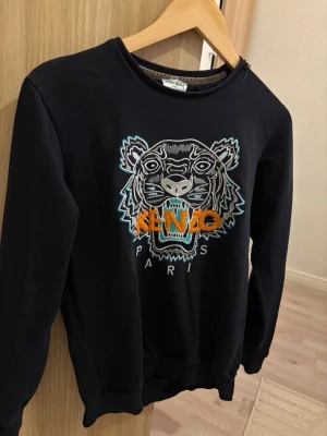 Svart Kenzo sweatshirt med tigertryck - Svart sweatshirt från Kenzo med ikoniskt broderat tigerhuvud och färgglad logga framtill. Rund halsringning, långa ärmar och ribbade muddar. Perfekt för dig som vill ha en statementtröja med streetwear-vibe.