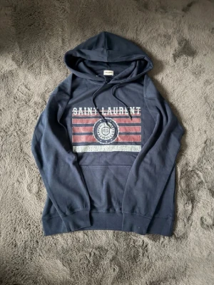 Saint Laurent hoodie - Snygg mörkblå hoodie från Saint Laurent k storlek L, passar dig som är mellan 180-190 beroende på passform. Perfekt för en avslappnad look. Mycket bra skick utan några defekter. Säljer då jag växt ut den, prefekt hoodie nu till hösten och vintern