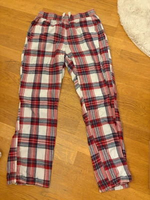 Rutig pyjamasbyxa i rött och vitt - Mysiga pyjamasbyxor med klassiskt rutigt mönster i rött, vitt och blått. Byxorna har resår i midjan och raka ben, perfekta för hemmakvällar. Tillverkade i mjukt tyg som känns skönt mot huden.