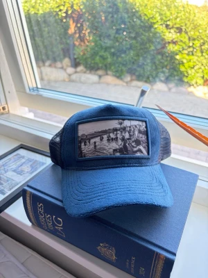 Blå truckerkeps med fototryck - Snygg blå truckerkeps med svart mesh baktill och ett stort svartvitt fototryck framtill som föreställer en strand- eller poolscen. Kepsen har böjd skärm och justerbar snapback. Perfekt för dig som vill sticka ut med en unik accessoar.