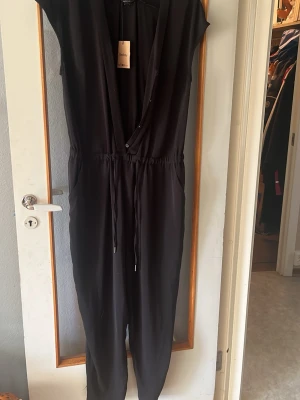 Svart jumpsuit med dragsko - Snygg svart jumpsuit med djup V-ringning och dragsko i midjan. Modellen har korta ärmar, långa ben och fickor på sidorna. Tillverkad i ett mjukt, tunt tyg som ger en avslappnad och trendig look.