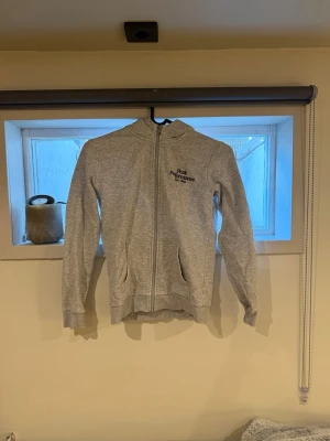 Grå zip hoodie från Peak Performance - Säljer en ljusgrå hoodie från Peak Performance med dragkedja framtill och två fickor. Tröjan har huva och ribbade muddar vid ärmslut och nederkant. Perfekt för en avslappnad och sportig stil.
