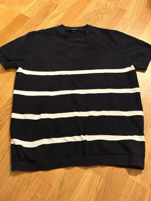 Randig t-shirt från Massimo Dutti - Snygg marinblå t-shirt från Massimo Dutti med vita horisontella ränder. Klassisk rund halsringning och korta ärmar. Tillverkad i exklusiv bomull som känns mjuk mot huden. Perfekt för en stilren och avslappnad look.