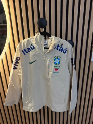 Vit Brasil Nike träningsjacka - Säljer en vit träningsjacka från Nike med Brasilien CBF-logga och sponsorer som Itaú och Guaraná. Jackan har huva, dragkedja framtill och tryck i blått, grönt och gult. Perfekt för dig som älskar fotboll och vill ha en unik sportig look.