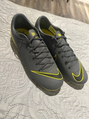 Gråa Nike fotbollsskor med dobbar - Snygga gråa fotbollsskor från Nike med gula detaljer och dobbar för bra grepp på planen. Skorna har snörning och en modern design med gula Nike-loggor och mönster på sulan. Perfekta för dig som vill sticka ut på fotbollsplanen.