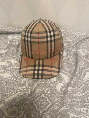 Beige keps med Burberry-rutor - Säljer en klassisk beige keps från Burberry med det ikoniska rutmönstret i svart, vitt och rött. Kepsen har böjd skärm och justerbar snapback baktill. Märkt med 'BURBERRY LONDON ENGLAND' på baksidan. Perfekt för dig som gillar stilrena accessoarer. Vid snabbaffär kan priset diskuteras. (Äkta kvitto finns) kan behövas tvätta kepsen där framme annars som helt ny!