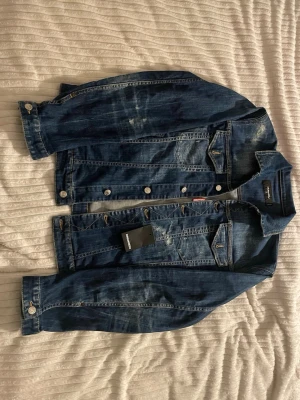 Mörkblå klassisk jeansjacka - Jackan är tillverkad i Italien och har den karakteristiska ICON-designen på ryggen, detaljerad denimtvätt och DSQUARED2-märkning. Mycket snygg och unik modell som passar perfekt för en mer exklusiv streetwear-look. Tillkommer även med få extra knappar. Inget kvitto. Säljs för den inte används. 4500 eller bud.
