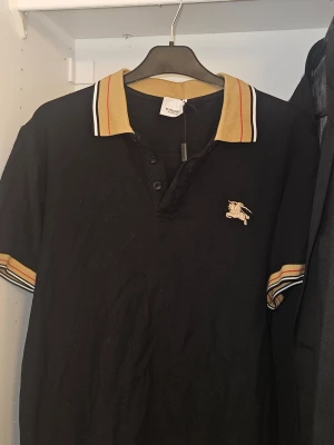 Svart burberry pikétröja  - Svart pikétröja med klassisk beige krage och ränder i rött och svart. Tröjan har korta ärmar med matchande detaljer och knappslå framtill. Broderad logga på bröstet ger en stilren touch. Perfekt för en avslappnad men snygg look.