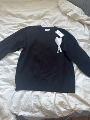 Svart stickad tröja från Ami - Svart stickad tröja från Ami med rund halsringning och ribbade muddar. På bröstet finns ett vitt hjärta med bokstaven 'A' broderat. Perfekt för dig som gillar stilrena plagg med en twist.