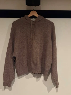 Brun stickad hoodie med huva - Mysig brun stickad hoodie med huva och dragsko. Tröjan har avslappnad passform, långa ärmar och ribbade muddar vid ärmslut och nederkant. Perfekt för chill dagar och enkel att matcha med jeans eller mjukisbyxor.
