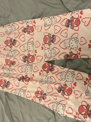 Hello Kitty & Spiderman pyjamasbyxor - Säljer ett par ljusrosa pyjamasbyxor med gulligt Hello Kitty och Spiderman-motiv samt röda hjärtan över hela tyget. Byxorna har rak passform och resår i midjan för extra komfort. Perfekta för mysiga hemmakvällar! Passar xs-m skulle jag vilja säga