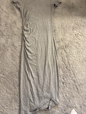 Grå ribbad kortklänning med rynk - Snygg grå kortklänning i ribbad bomullsmix med rynkdetalj på sidan. Klänningen har korta ärmar och en enkel, figurnära passform som ger en stilren look. Perfekt för dig som gillar minimalistisk stil med en twist.