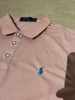 Rosa pikétröja från Polo Ralph Lauren - Snygg rosa pikétröja från Polo Ralph Lauren med klassisk krage, tre knappar och blå broderad logga på bröstet. Perfekt för en stilren och avslappnad look. Tillverkad i mjuk bomull som känns skön mot huden.