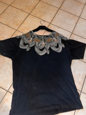 Svart t-shirt Marcelo Burlon med mönster - Säljer en svart t-shirt från Marcelo Burlon med ett grått och brunt grafiskt mönster runt halsen. T-shirten har korta ärmar och en normal passform. Perfekt för dig som vill sticka ut med en unik design.