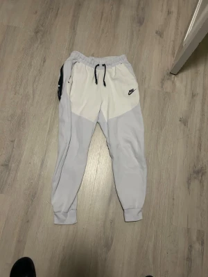 Niketech vita mjukisbyxor med snörning - Niketech i vitt med svart snörning i midjan och logga på vänster sida. Byxorna har muddar vid bensluten och en kontrastfärgad panel på sidan. Perfekta för chill dagar eller träning. En stor och rymlig ficka finns på sidan med dragkedja. Säljes pågrund av lite användning och tyvärr ett litet hål vid fickan.