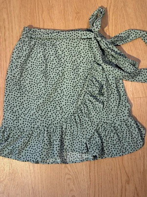 Grön prickig omlottkjol med volang - Söt grön kort omlottkjol med svarta prickar och volangkant. Kjolen har knytband i midjan och är gjord i ett lätt, luftigt material som passar perfekt till varmare dagar. Den har en avslappnad och trendig look.