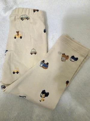 Beige leggings med bilar från Lindex - Söta beige leggings från Lindex med gulligt mönster av små bilar, helikoptrar och björnar. Mjukt och stretchigt material med resår i midjan för skön passform. Perfekta för en lekfull vardagsstil till de minsta. Fått I gåva utan Tag. Storlek 80