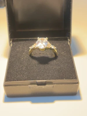 Guld ring - Snygg guldfärgad ring med en stor, klar oval sten i mitten och mindre stenar på sidorna. Ringen har en klassisk och elegant design som verkligen fångar blicken. Perfekt för dig som vill ha något som sticker ut och känns lyxigt. Storlek 4,5