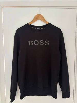Svart sweatshirt från BOSS - Stilren svart sweatshirt från BOSS med rund halsringning och broderad logga på bröstet. Tröjan har långa ärmar, ribbade muddar och är tillverkad i mjukt material som passar perfekt till en avslappnad look.