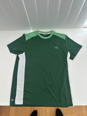 Grön Lacoste t-shirt med vita detaljer sällsynt!!  - Snygg grön t-shirt från Lacoste med ljusgröna axelpartier och vita sidopaneler. Klassisk Lacoste-logga på bröstet och stor logotyp med krokodil på ryggen. Tillverkad i ett snabbtorkande polyestermaterial, perfekt för sportiga aktiviteter. Äkta 