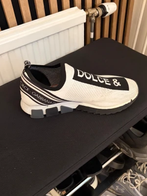 Dolce & Gabbana vita slip-on sneakers - Stilrena vita slip-on sneakers från Dolce & Gabbana med svart och vit logga på ovandelen. Skorna har en stickad meshstruktur, elastisk öppning och chunky sula med grå detaljer. Perfekta för dig som vill ha en sportig och trendig look.