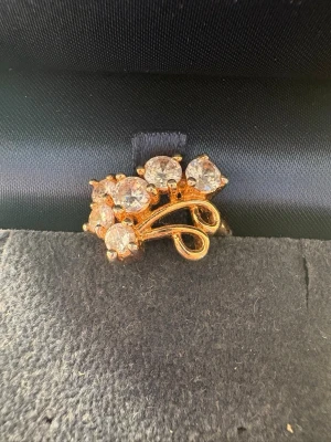 Guldfärgad ring med stora stenar - Unik guldfärgad ring med sex stora, gnistrande stenar i klusterdesign. Ringen har en elegant, snirklig form som ger ett lyxigt intryck och passar perfekt för dig som gillar statement-smycken. Materialet ser ut som metall med glittrande stenar.