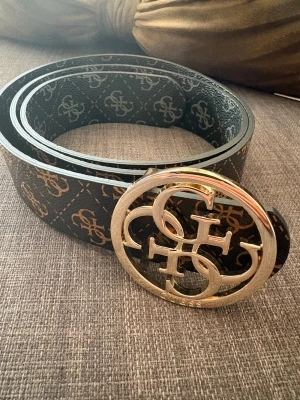 Svart bälte med guldspänne och logga - Snyggt svart bälte med guldfärgat stort spänne och mönster av loggor längs hela bandet. Perfekt accessoar för att lyfta din outfit med en exklusiv touch. Bältet har en klassisk form och detaljer i guld.
