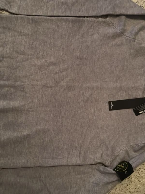 Grå stickad tröja från Stone Island - Snygg grå stickad tröja från Stone Island med klassisk logotyp-patch på ärmen. Tröjan har rund halsringning och ribbade muddar vid ärmslut och nederkant. Perfekt för dig som gillar stilrena och trendiga plagg.