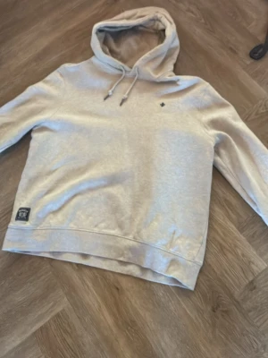 Beige hoodie med patch och brodyr - Säljer en beige hoodie med dragsko i huvan och liten broderad logga på bröstet. Nedtill finns en patch med text och motiv. Hoodien har ribbade muddar och är tillverkad i mjukt sweatshirtmaterial. Perfekt för en avslappnad och stilren look.