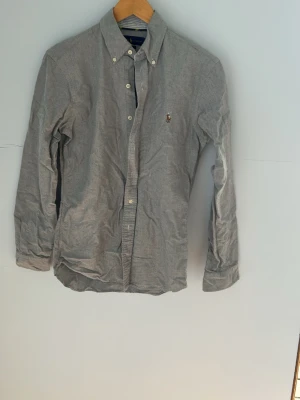 Grå skjorta från Polo Ralph Lauren - Klassisk grå skjorta från Polo Ralph Lauren med broderad logga på bröstet. Skjortan har knappar framtill, button-down krage och långa ärmar. Perfekt till både jeans och chinos för en stilren look.