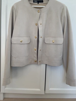Beige mockaliknande jacka från Zara - Stilren beige jacka från Zara i mockaliknande material. Jackan har rund halsringning, guldknappar framtill och två stora fickor med lock. Croppad modell med lång ärm, perfekt för en trendig look.